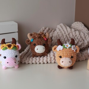 May include: Three crocheted cow plushies in white, brown, and orange, each with horns and floral headbands. The plushies are on a white surface with a cream-colored blanket in the background. A wooden box with white yarn is on the right.