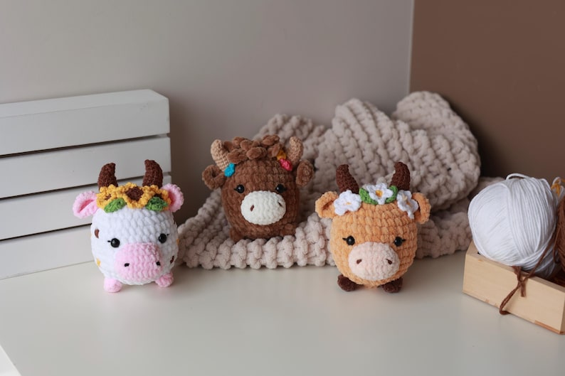 May include: Three crocheted cow plushies in white, brown, and orange, each with horns and floral headbands. The plushies are on a white surface with a cream-colored blanket in the background. A wooden box with white yarn is on the right.
