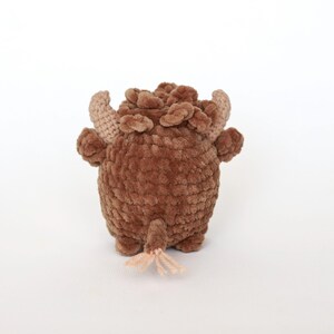 May include: A handmade brown crochet bull toy with tan horns and a small tail. The plush toy has a textured, fuzzy appearance and is standing on a white surface. The bull has small, rounded feet and a detailed head.