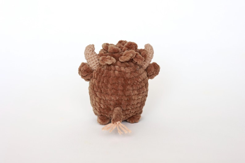 May include: A handmade brown crochet bull toy with tan horns and a small tail. The plush toy has a textured, fuzzy appearance and is standing on a white surface. The bull has small, rounded feet and a detailed head.
