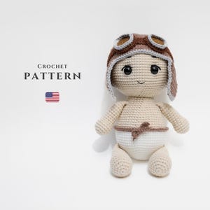 May include: A crocheted doll wearing a brown aviator hat with goggles and a white diaper. The doll is beige and has black eyes and a small smile. The text "CROCHET PATTERN" is above the American flag.