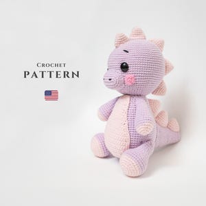 Könnte beinhalten: Ein gehäkeltes lila und rosa Dinosaurierspielzeug mit schwarzen Augen und rosa Wangen. Das Spielzeug sitzt auf einer weißen Oberfläche. CROCHET PATTERN steht in schwarzer Schrift über dem Spielzeug.