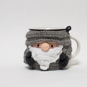 May include: A gray crocheted mug cozy shaped like a gnome with a white beard and a black hat. The cozy is on a white mug.