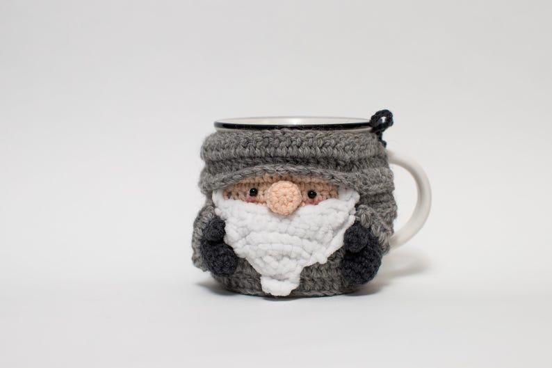 May include: A gray crocheted mug cozy shaped like a gnome with a white beard and a black hat. The cozy is on a white mug.