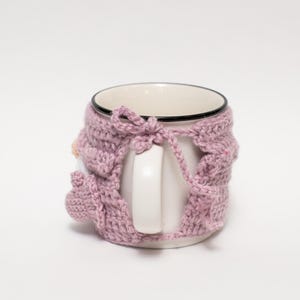 May include: A white ceramic mug with a pink crocheted cozy. The cozy has a bow at the top and covers the entire mug, including the handle.