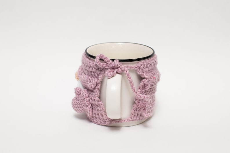 May include: A white ceramic mug with a pink crocheted cozy. The cozy has a bow at the top and covers the entire mug, including the handle.