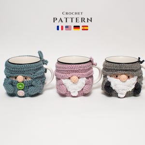 May include: Three crocheted mug cozies in the shape of gnomes. The cozies are in blue, pink, and grey. The cozies have white beards and black eyes. The text "CROCHET PATTERN" is above the cozies.