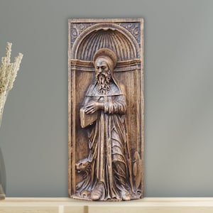 May include: A wooden wall plaque with a carved relief of a bearded man in a robe, holding a book. The plaque is framed with ornate carvings and has a brown finish.