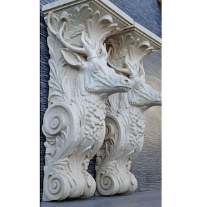 May include: Two off-white, carved deer head corbels. The deer heads have detailed antlers and are surrounded by ornate, swirling leaf designs. The corbels are mounted on a dark gray wall.