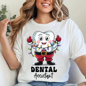 May include: A white t-shirt featuring a cartoon tooth character dressed as Santa Claus, wearing red pants, a black belt with a gold buckle, and red gloves. The tooth is tangled in colorful Christmas lights. The text DENTAL ASSISTANT is printed below.