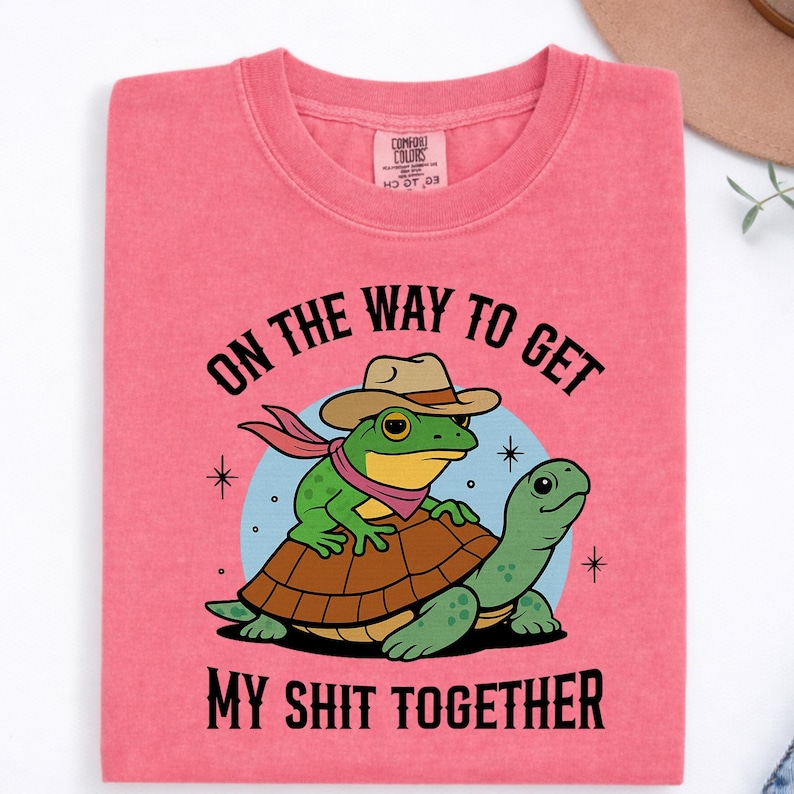May include: A coral pink t-shirt featuring a graphic of a green frog wearing a tan cowboy hat and a pink scarf, riding on the back of a green turtle. The text reads ON THE WAY TO GET MY SHIT TOGETHER. The shirt has a Comfort Colors brand label.