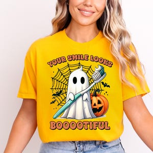 May include: A bright yellow t-shirt featuring a graphic of a white ghost holding a toothbrush with toothpaste, a jack-o'-lantern, and a spiderweb. The text reads "YOUR SMILE LOOKS BOOOOTIFUL" in orange letters with a dark outline.
