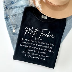 Puede incluir: Camiseta negra con la frase "Math Teacher" en cursiva. La camiseta incluye una definición de profesor de matemáticas. La camiseta está colocada sobre unos vaqueros azules y junto a un sombrero beige.