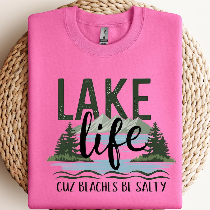 May include: A bright pink crewneck sweatshirt featuring a graphic print with the text "LAKE life" in dark green and black, accompanied by an illustration of mountains, pine trees, and water, with the text "CUZ BEACHES BE SALTY" below.