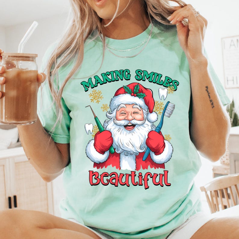 May include: A light mint green t-shirt featuring a graphic of Santa Claus wearing a red hat, holding a green dental pick and a blue toothbrush. The text reads Making Smiles Beautiful in green and red, surrounded by small white tooth icons.