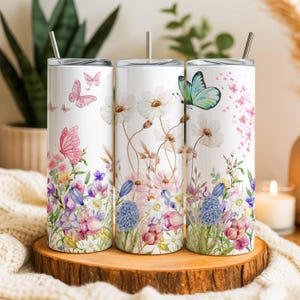 May include: Three white stainless steel tumblers with floral and butterfly designs. The tumblers feature watercolour illustrations of pink, purple, and blue flowers, with butterflies in pink and teal. Each tumbler has a silver straw.
