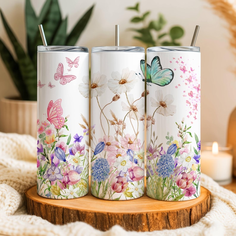May include: Three white stainless steel tumblers with floral and butterfly designs. The tumblers feature watercolor illustrations of pink, purple, and blue flowers, with butterflies in shades of pink and teal. Each tumbler has a silver straw.