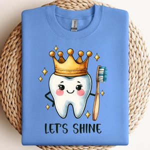 May include: A light blue crewneck sweatshirt featuring a cartoon tooth wearing a gold crown and holding a toothbrush. The tooth has a smiling face with pink cheeks and is surrounded by small yellow stars. The text LET'S SHINE is printed below.