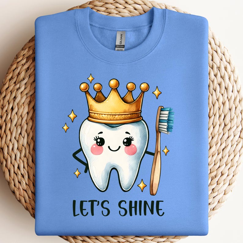 May include: A light blue crewneck sweatshirt featuring a cartoon tooth wearing a gold crown and holding a toothbrush. The tooth has a smiling face with pink cheeks and is surrounded by small yellow stars. The text LET'S SHINE is printed below.