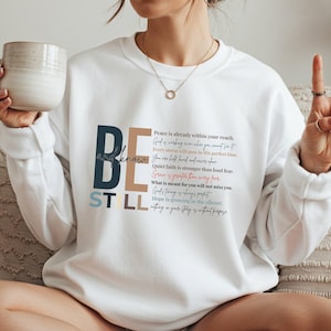 Può includere: Felpa bianca con le parole "BE STILL and know" in blu, marrone e beige. La felpa presenta anche un elenco di citazioni ispiratrici in una miscela di colori. Uno stile casual e confortevole.