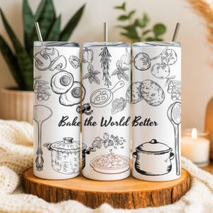 May include: Three white stainless steel insulated tumblers with black line art illustrations of cooking ingredients and utensils. The text "Bake the World Better" is printed on the tumblers. Each tumbler has a silver straw.