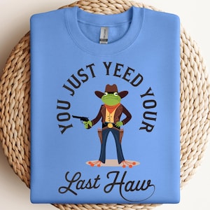 May include: A light blue crewneck sweatshirt featuring a graphic of a green frog dressed as a cowboy, wearing a brown hat, jacket, and holster. The text reads, "YOU JUST YEED YOUR LAST HAW" in black lettering surrounding the frog illustration.