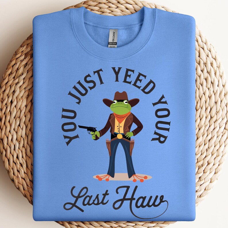May include: A light blue crewneck sweatshirt featuring a graphic of a green frog dressed as a cowboy, wearing a brown hat, jacket, and holster. The text reads, "YOU JUST YEED YOUR LAST HAW" in black lettering surrounding the frog illustration.