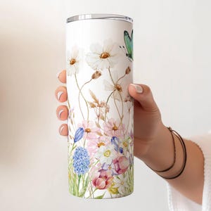 May include: A white tumbler featuring a watercolor floral design with white flowers, pink and blue blooms, and a green butterfly. The tumbler is held by a person's hand.