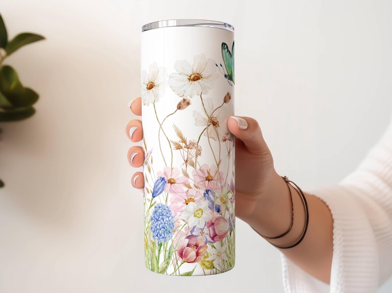 May include: A white tumbler featuring a watercolor floral design with white flowers, pink and blue blooms, and a green butterfly. The tumbler is held by a person's hand.