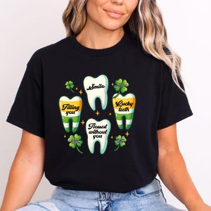 May include: A black t-shirt featuring a graphic print of four white teeth arranged in a circle. The teeth contain the text "Smile", "Filling you", "Lucky teeth", and "Flossed without you". Green four-leaf clovers and small yellow stars surround the teeth.