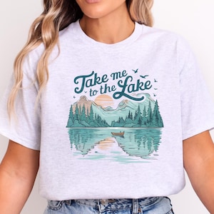 May include: A light gray heathered t-shirt featuring a graphic print of a mountain lake scene with a small boat, pine trees, and a sunset. The text "Take me to the Lake" is printed in dark teal script above the landscape, accompanied by small birds.
