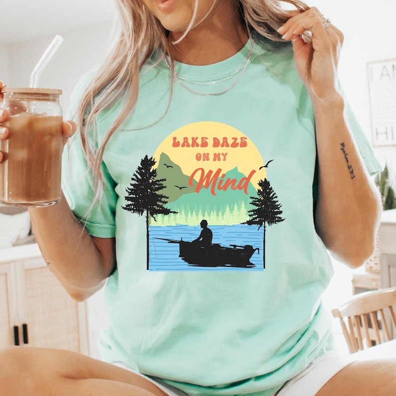 May include: A light mint green t-shirt featuring a graphic of a person fishing in a boat on a lake. The design includes a yellow sun, green mountains, black pine trees, and the text "LAKE DAZE ON MY MIND" in orange and brown lettering.