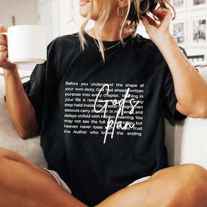 Può includere: T-shirt nero con testo bianco "God's plan" in un carattere scritto a mano. La maglietta ha anche un paragrafo di testo più lungo in un carattere più piccolo. La maglietta è in stile casual.