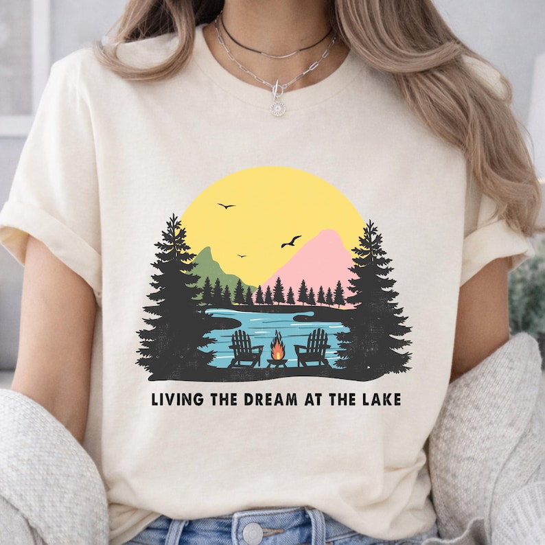 May include: A cream-colored short-sleeve t-shirt featuring a graphic print of a lake scene with two chairs, a campfire, pine trees, mountains, and a yellow sun. The text "LIVING THE DREAM AT THE LAKE" is printed in black below the graphic.