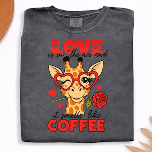 May include: A charcoal grey t-shirt featuring a graphic of a giraffe wearing red heart-shaped glasses, winking, and holding a red rose in its mouth. The text reads LOVE is in the air and it smells like COFFEE, surrounded by small red hearts.