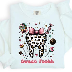 May include: A white long-sleeve shirt featuring a central graphic of a tooth with a leopard print pattern and a pink bow on top. The design is surrounded by colorful candies, lollipops, and small hearts, with the text Sweet Tooth in pink below.