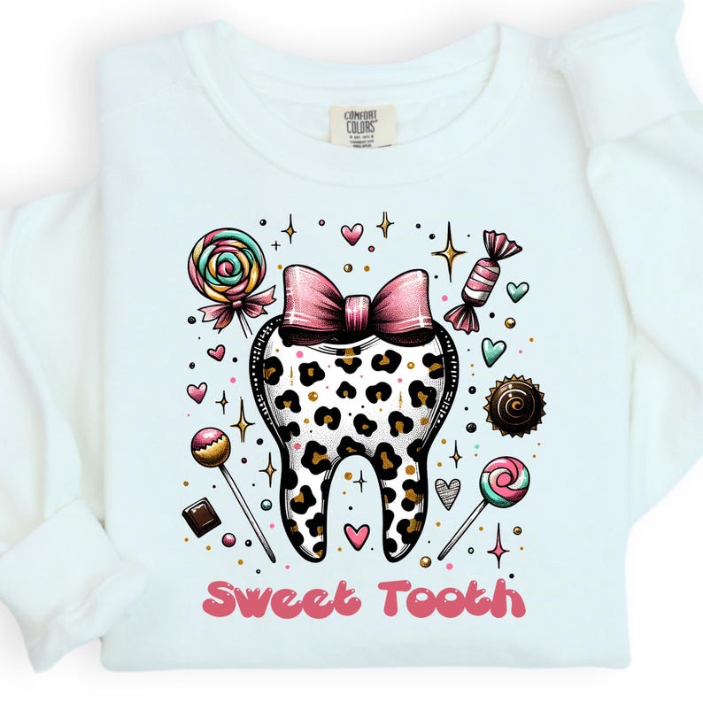 May include: A white long-sleeve shirt featuring a central graphic of a tooth with a leopard print pattern and a pink bow on top. The design is surrounded by colorful candies, lollipops, and small hearts, with the text Sweet Tooth in pink below.