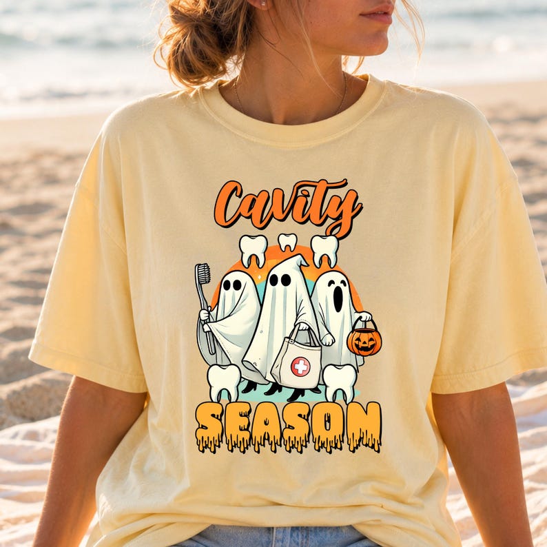 May include: A pale yellow t-shirt featuring a graphic of three ghosts dressed in white sheets. The ghosts are holding a toothbrush, a medical bag with a red cross, and a jack-o'-lantern bucket. The text reads Cavity SEASON with teeth icons.