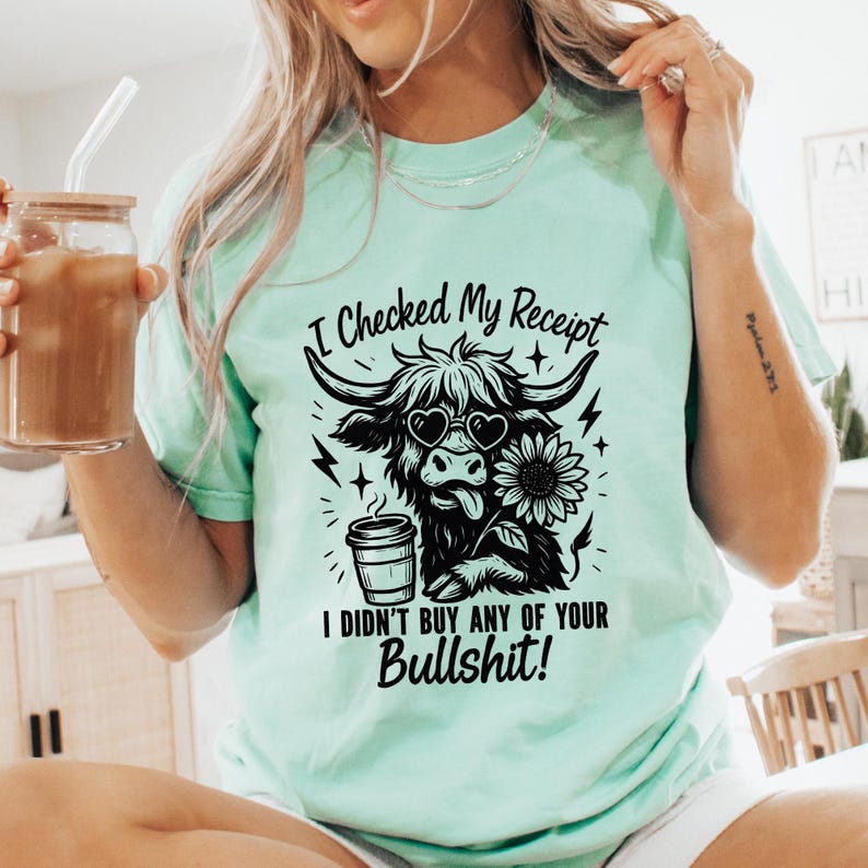 May include: A mint green short-sleeve t-shirt featuring a black graphic of a Highland cow wearing heart-shaped sunglasses, holding a coffee cup, and next to a sunflower. The text reads: I Checked My Receipt I DIDN'T BUY ANY OF YOUR BULLSHIT!
