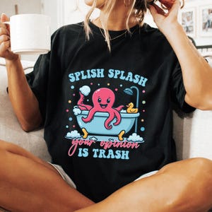 May include: A black t-shirt featuring a graphic of a pink octopus in a light blue bathtub with a yellow rubber duck, bubbles, and a shower head. The text reads "SPLISH SPLASH" in light blue and "your opinion IS TRASH" in pink and light blue.
