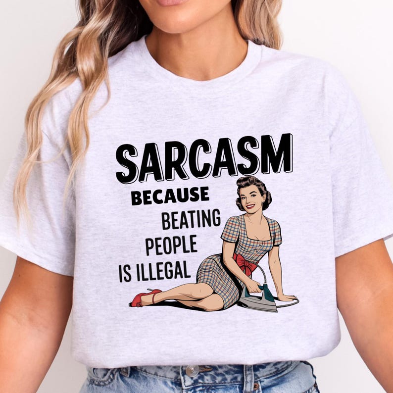 May include: A light grey short-sleeved t-shirt featuring a graphic of a person in a plaid dress with a red bow and a clothing iron. The text reads: SARCASM BECAUSE BEATING PEOPLE IS ILLEGAL. The design is printed in black, red, and blue ink.