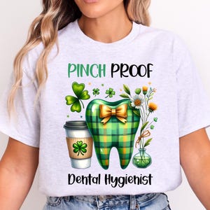 May include: A light gray t-shirt featuring a graphic of a green plaid tooth with a gold bow, a coffee cup with a shamrock, and a small vase of flowers. The text reads PINCH PROOF and DENTAL HYGIENIST in black and green lettering.
