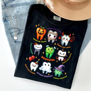 May include: A black t-shirt featuring a graphic of nine cartoon teeth dressed in Halloween costumes, including a pumpkin, mummy, devil, skeleton, Frankenstein, vampire, party guest, ghost, and witch, with text including Trick or Treat and Bootiful.