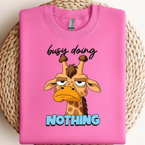 May include: A bright pink crewneck sweatshirt featuring a cartoon giraffe with a bored expression. Above the giraffe, the text reads busy doing in black script, and below the giraffe, the word NOTHING is printed in bold light blue block letters.