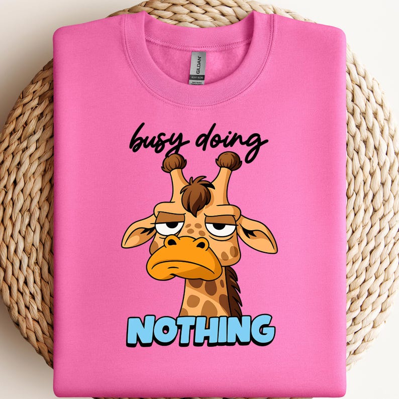 May include: A bright pink crewneck sweatshirt featuring a cartoon giraffe with a bored expression. Above the giraffe, the text reads busy doing in black script, and below the giraffe, the word NOTHING is printed in bold light blue block letters.