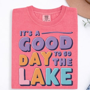May include: A coral pink crew neck t-shirt featuring the text IT'S A GOOD DAY TO GO THE LAKE in a multi-colored graphic. The words are printed in purple, orange, and light blue with a dark purple drop shadow. The shirt has a Comfort Colors label.