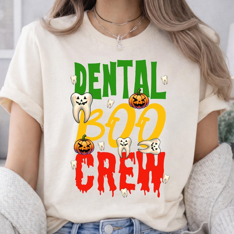 May include: A cream-colored short-sleeve t-shirt featuring the text DENTAL BOO CREW in green, yellow, and red. The design includes illustrations of teeth with jack-o'-lantern faces and orange pumpkins, creating a festive Halloween-themed graphic.