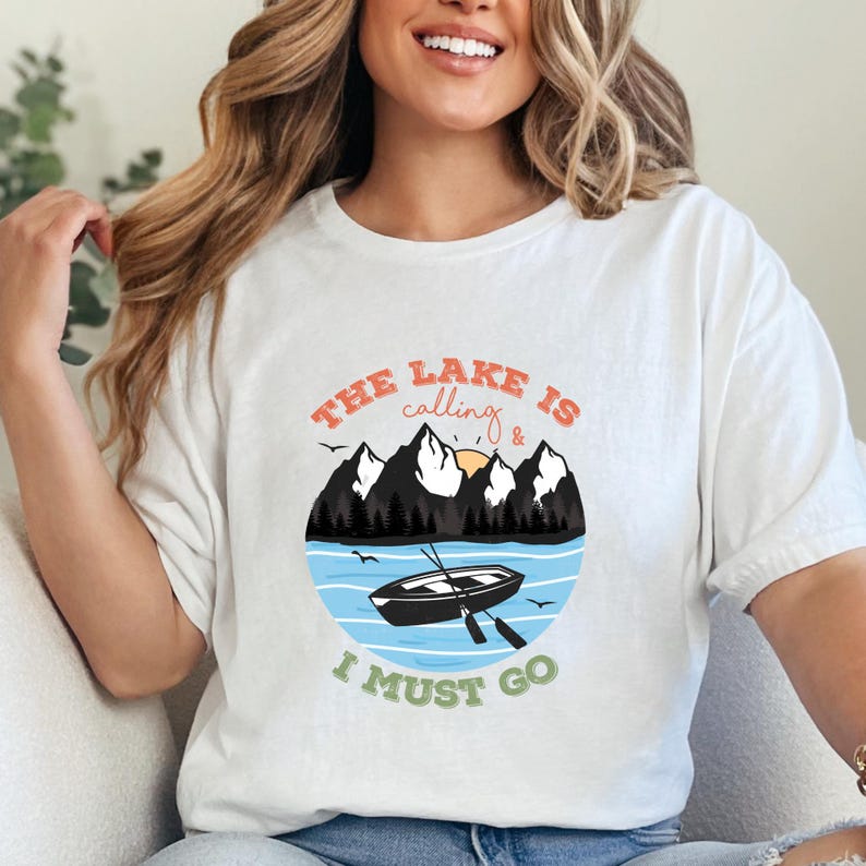 May include: A white short-sleeved t-shirt featuring a circular graphic of a black rowboat on blue water, dark mountains, and a yellow sun. The text reads THE LAKE IS calling & I MUST GO in orange, black, and green lettering across the design.