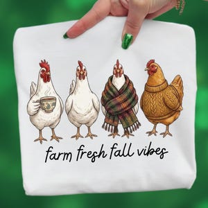 May include: A white t-shirt featuring a graphic of four chickens. One holds a coffee cup, one is plain, one wears a plaid scarf, and one wears a brown cable-knit sweater. Below the chickens, the text reads farm fresh fall vibes in black.