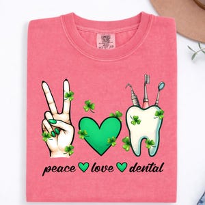 May include: A coral pink t-shirt featuring a graphic with a hand making a peace sign, a green heart, and a white tooth with dental tools, all decorated with green shamrocks. The text below reads peace, love, and dental with small heart icons.